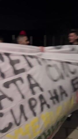 The video shows a group of men walking toward a stone wall and gate at night. Several men are carrying a banner with Cyrillic writing on it. The men approach the gate, and some climb over or go through it. The gate is made of metal and has an ornamental design. The wall is constructed of stone and has a fence on top. The environment appears to be residential, with street lights visible. The video is somewhat dark and shaky.