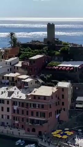 The video shows an aerial view of a coastal town. The town is characterized by colorful buildings clustered together, with a harbor filled with small boats. A stone tower is visible on a hill overlooking the town. The sea is calm and sparkles in the sunlight under a clear blue sky. People can be seen walking along the waterfront.
