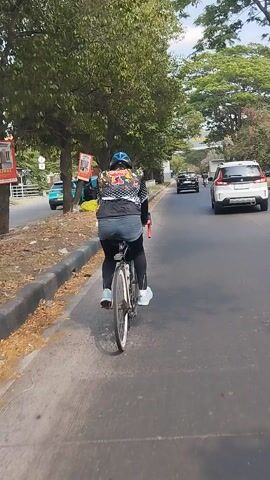 This video shows a person riding a bicycle on a paved road. The rider is wearing a black and white patterned jacket, black pants, a blue helmet, and white shoes. The bicycle has a water bottle cage and a kickstand. The scene is a roadside with trees and bushes on the left side. In the background, there are vehicles on the road, including a white car and a black car. There is also a green car parked on the side of the road. The time of day appears to be daytime, with clear skies and sunlight. The overall atmosphere is one of an outdoor activity. There is a sign with text on it visible near the bottom left of the frame, but the text is not legible. The video is oriented vertically.