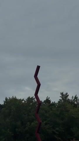 A red, zigzagging sculpture stands tall in an outdoor setting. The sculpture is made of metal and has a matte finish. It is placed in a grassy area with trees in the background. The sky is cloudy and overcast. The camera is positioned at a low angle, looking up at the sculpture. The video pans slowly upwards, revealing the entire sculpture.