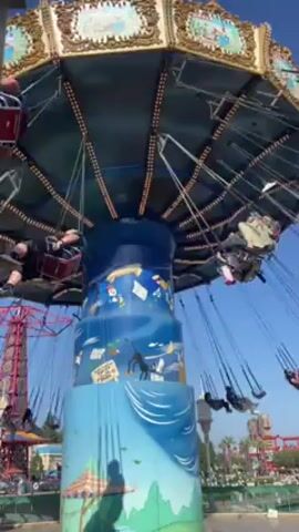 The video shows a spinning swing ride in an outdoor setting. The ride has a tall central column decorated with colorful illustrations. Around the top of the column, swings are suspended by chains, carrying riders in a circular motion. In the background, there are other structures and landscaping, all under a clear blue sky.
