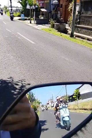The video captures a point-of-view shot from a motorcycle, showing a road with several other motorcycles and people. The road appears to be in a tropical or subtropical region, with lush greenery and traditional-style buildings lining the sides. The sky is clear and blue, suggesting a sunny day. The motorcycle is in motion, and the camera pans slightly, showing the rider's hand holding the handlebar and the side-view mirror. The mirror reflects a continuation of the road scene, with more motorcycles and pedestrians visible. The overall impression is of a busy street in a populated area, possibly a village or town.