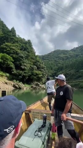 A video shows two men in a boat on a river. The man in the back is using a long pole to propel the boat through the water. The man in the front is smiling and waving at the camera. The boat is surrounded by lush green trees and mountains. The sky is cloudy.
