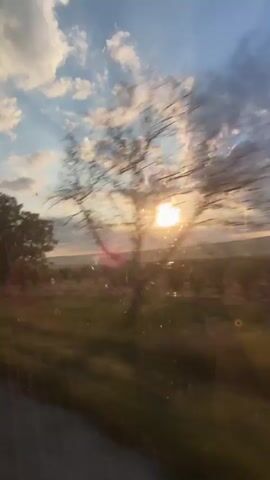 This is a video taken from inside a vehicle looking out a window at a rural landscape during sunset. The sun is visible, partially obscured by clouds and trees. The camera is moving, causing a slight blur in the foreground and some of the trees. The landscape appears to be fields with some trees. The sky is blue with scattered white and gray clouds. There are some light flares or water droplets on the window.