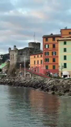 The video pans across a harbor landscape at dawn or dusk. Initially, the camera focuses on a castle-like structure on a rocky coastline, adjacent to a building with red and yellow walls. The camera then moves to show numerous boats anchored in the calm water. As the video progresses, the sky transitions into a vibrant gradient of oranges and yellows as the sun sets or rises, casting a warm glow on the water and surrounding landscape. Mountains are visible on the horizon under the colorful sky, adding depth to the view. The overall mood is peaceful and serene.