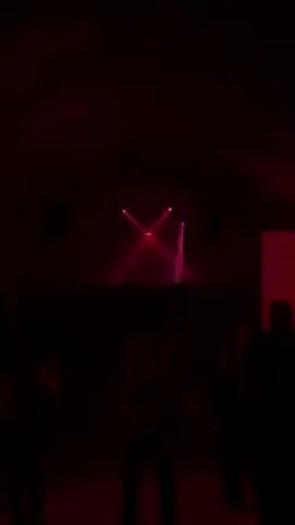 The scene is a dark, possibly indoor space with several people visible as silhouettes. A red light is prominent in the center and is accompanied by moving red laser beams that sweep across the space. Speakers are visible on the walls. The overall atmosphere suggests a club or event with dynamic lighting effects.