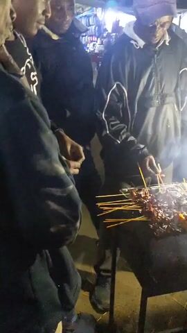 Here is a detailed description of the video:

The video shows a nighttime scene of a street food vendor grilling meat skewers. The vendor, wearing a dark jacket and a baseball cap, is tending to the grill, which is emitting a significant amount of smoke. Several people are standing around the grill, presumably waiting to purchase the skewers. The grill itself is a simple metal structure with a grate on top, under which red-hot coals are visible. Several skewers with pieces of meat on them are cooking on the grill. A variety of condiments, including a red sauce in a plastic container and a green liquid in a cup, are visible on a nearby table. The background is blurred, but it appears to be a busy street with cars and people passing by.