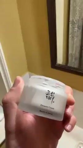 A person holds a jar of Beauty of Joseon Dynasty Cream. They place the jar on a shelf with a white towel, deodorant, and toothpaste.  The person takes a selfie in the mirror while applying the cream to their face. They are wearing a navy blue striped shirt. The bathroom has yellow walls and a framed mirror.
