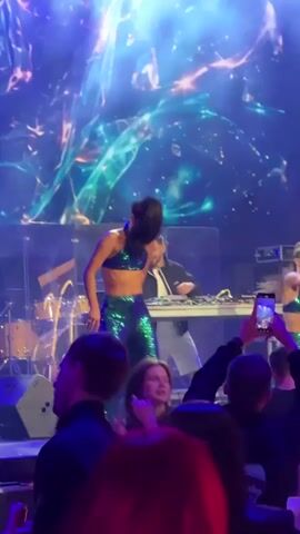 A short video clip shows a female dancer in a sparkly green outfit performing on a stage with a DJ in the background. The dancer is center frame, wearing a crop top and wide-leg pants, with her hair pulled back. She is raising one arm in the air and appears to be singing or interacting with the audience. Behind her, a male DJ is seated at a console, and another female is partially visible to his right. The stage is lit with blue and purple lights, and a large, abstract visual projection is displayed behind the performers. In the foreground, audience members are visible, many holding up their phones to record the performance. The scene captures a moment of energetic live entertainment.