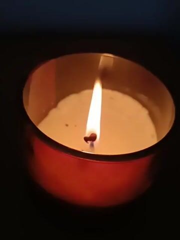 A close-up shot captures a lit candle in a red container. The flame flickers gently against a dark background. The candle wax appears creamy white with a few dark specks.