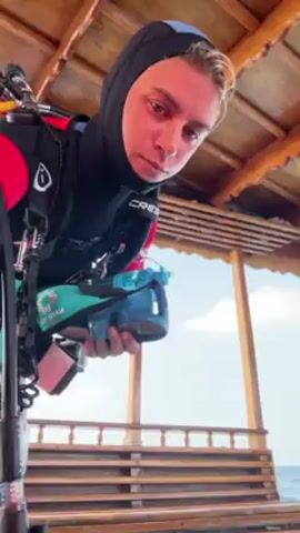 Here is a description of the video:
A person wearing a full scuba suit and hood is standing under a wooden structure. The person is holding a pair of blue and white scuba diving goggles in their hands and spraying them with a clear liquid from a white spray bottle. The person is standing on a wooden platform with the ocean visible in the background. The wooden structure has a ceiling made of wooden planks.