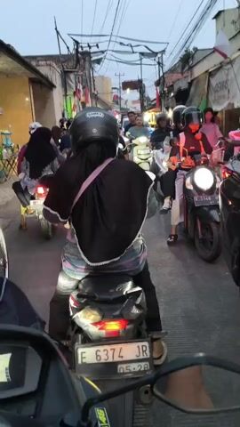 A first-person perspective shot from the back of a motorcycle shows a congested street. Many motorcycles are present, with riders wearing helmets and some with passengers. A sign with "Zhafirah" is visible hanging above the street, along with numerous electrical wires and cables. Indonesian flags are also displayed on some buildings. The lighting suggests it is either early morning or late evening. The motorcycles are stopped, indicating traffic congestion.
