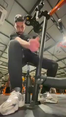 The video shows a person sitting on a workout machine in a gym and working out. The person is wearing a black shirt, gray sweatpants, and white and gray sneakers. They are holding onto the machine's handles and extending their arms outward. The machine is black and orange. The gym has a high ceiling with exposed metal beams and rows of other workout machines.