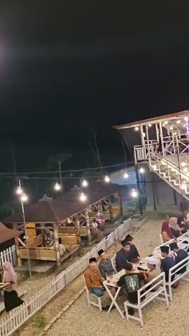 The video shows an outdoor gathering at night. The scene is lit by string lights and a few standing lamps. There are multiple seating areas, including picnic tables and individual tables with chairs. Some people are seated at the tables, appearing to be eating or socializing. There is a two-story structure in the background with more lights and people on the upper level. Several motorcycles are parked to the side. The ground is covered in gravel. The overall atmosphere appears to be relaxed and casual.