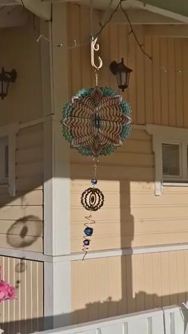 The video shows a decorative wind spinner hanging from a porch roof. The spinner is made of polished metal with a patterned design and is adorned with blue glass beads. It is suspended by a chain and hook, with a string of small fairy lights draped above it. The background consists of the beige wooden siding of a house with white trim. A black outdoor lantern is mounted on the wall. Shadows of the spinner and its surroundings are cast on the wall. The overall scene is a close-up view of the hanging ornament on a bright, sunny day.
