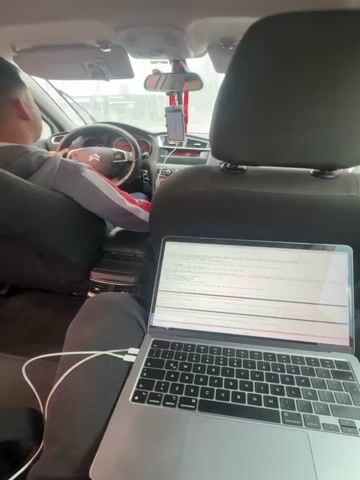 The video shows the interior of a car on a rainy day. A driver is visible in the front seat, focused on driving. A passenger is in the back seat using a silver MacBook with a white charging cable.  Rain is visible on the window. Buildings and cars can be seen outside.