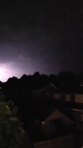 A nighttime video captures a residential area, likely suburban. The scene is dark, with the ambient light emanating from streetlights and the illuminated sky. Houses and trees are visible in silhouette against the dark sky. The upper portion of the video shows a bright flash of light, which appears to be lightning, illuminating the clouds with a purple and white glow.