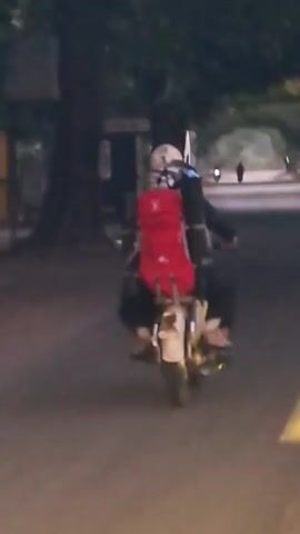 A person wearing a helmet and a large red backpack is riding a motorcycle on a paved road at night. They are moving away from the camera. There are trees and a building on the left side of the road. On the right side of the road, there are several buses parked. The road has yellow lane markings.
