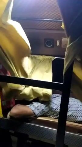 The video shows the interior of what appears to be a sleeper bus. A person is lying down on the lower bunk, covered with a patterned blanket or sheet. The bunk is made of a metal frame. Above the bunk are yellow curtains that are partially drawn, obscuring the upper bunk. The ceiling of the bus has a patterned design. There is a hanging garment and some bags visible next to the bunk.