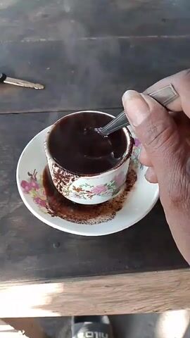 A close-up shot shows a hand holding a spoon and stirring a cup of dark coffee. The coffee is in a white cup with a floral pattern, resting on a saucer. Coffee grounds are spilled on the saucer. In the background, a wooden surface is visible, with a set of keys lying on it. The overall lighting suggests an outdoor or dimly lit indoor setting.