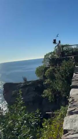 The video shows a bright, sunny day at the edge of a rocky cliff overlooking a calm ocean. The camera pans from a closer view of the cliff edge with some shrubbery to a wider shot of the ocean, showing the sun's reflection shimmering on the water. In the distance, the coastline is visible with hills covered in greenery. The sky is clear and blue, and the overall scene is peaceful and scenic.