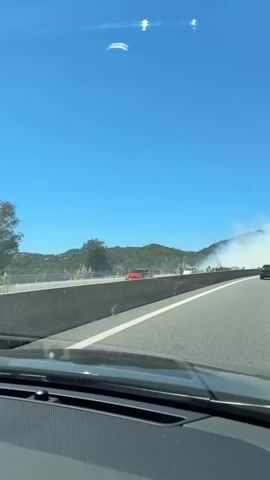 The video captures a view from inside a vehicle driving on a highway. A bright blue sky is visible overhead. To the left of the road, beyond a dark mesh barrier, is a landscape with trees and hills. A plume of white smoke is rising from the vegetation on the left side of the road, obscuring some of the trees and hills. A dark-colored car is seen in the smoke on the left side of the highway. Further down the road, the smoke appears to be spreading and becoming denser, creating a hazy atmosphere. The road itself has a white line marking the lane. The view is slightly obscured by the dashboard of the vehicle.