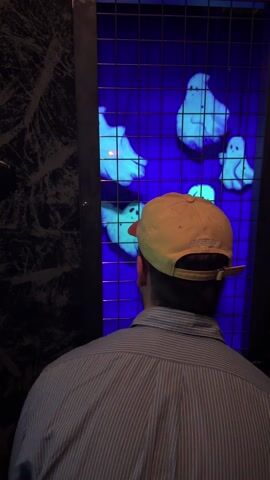 A person in a striped shirt enters a dark room and picks up a black gun-like object. They stand in front of a screen with glowing blue lights and aim at the screen. The screen displays a grid and glowing figures. A yellow baseball cap appears later, worn by another person also standing in front of the screen. A monitor displays a 52% score.