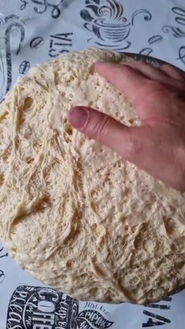 A hand presses into a large ball of risen dough that is in a round pan. The dough is light in color and has a very airy and bubbly texture. The pan sits on a surface covered with a white cloth that has black line drawings of coffee cups and coffee beans printed on it. The hand is seen from an overhead perspective, and the fingers are partially submerged into the dough. The camera moves slightly throughout the short video, zooming in and out and shifting focus.