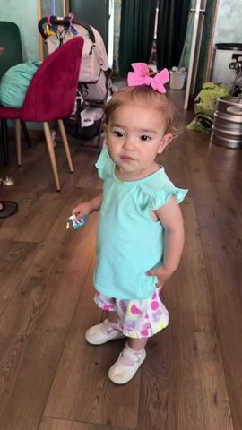 A toddler stands on a wooden floor, glancing around. The child has curly, light brown hair tied up with a pink bow. They wear a light blue t-shirt with ruffled sleeves, patterned shorts with pink, white, and purple designs, and white shoes with a strap. In their left hand, they hold a small, wrapped candy. Their right hand is tucked into their short's pocket. The background includes a red chair with a green cushion, a stroller, green curtains, and a metal keg. A pair of shoes are visible under the chair.