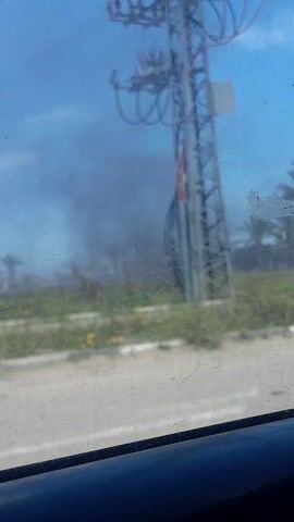 The video shows smoke rising in the distance beyond a road. A utility tower is visible on the left side of the frame. Palm trees and some vegetation are visible in the background. A white van passes by. The sky is blue. The video is taken from inside a moving vehicle.