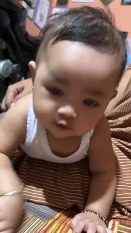 The video shows a baby lying on its stomach on a patterned surface. The baby is wearing a white tank top and a brown and white striped skirt. The baby's face is visible, and it is looking towards the camera. In the background, there are some objects, including what appears to be a fan and some framed pictures on the wall. A person's arm is visible holding the baby. The scene appears to be indoors, in a domestic setting. The lighting is warm and somewhat dim. The baby is the primary focus of the video.
