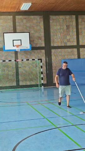 A man in a dark blue t-shirt and light-colored shorts is mopping a blue gymnasium floor. He is holding a mop with a white handle and appears to be moving from right to left across the frame. The gymnasium has a wooden ceiling, brick walls, and a basketball hoop on the left side. There is also a green and black netted soccer goal in the background.