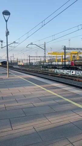 The video shows a train with the Rodalies logo and the Renfe logo passing by a train station platform. The train is white with orange and purple accents. It is moving from right to left in the frame. The platform is paved with grey tiles and has a yellow safety line along the edge. There are train tracks, overhead power lines, and streetlights visible. The sky is clear and blue, suggesting it is daytime. The scene appears to be a typical train station environment.