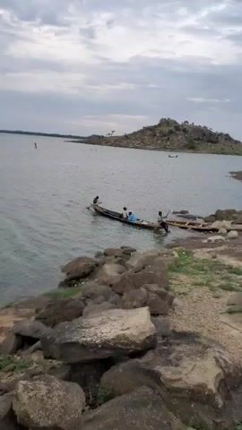 A narrow wooden boat with three people is moored to a wooden jetty on the shore of a large body of water. Rocks and sparse vegetation are visible along the shoreline. In the distance, a rocky island rises from the water, and the sky is overcast. The overall atmosphere is calm and serene.