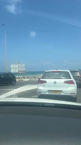 A white Volkswagen Golf is stopped at a traffic light on a sunny day. The car is in the foreground, and the ocean is visible in the background. There are other cars around, but they are not as clear. The sky is blue with a few clouds. There are also some signs and a lamppost.