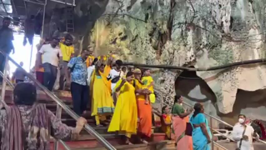 People are walking down a staircase in a cave. The staircase has metal railings. The people appear to be of South Asian descent. Many are wearing yellow clothing. Some people are raising their hands in the air. The cave walls are visible behind the people on the stairs and are a mixture of dark and light colors, with some holes and crevices. There are also some metal pipes and wires visible on the cave ceiling. Some people on the stairs are looking at their phones.
