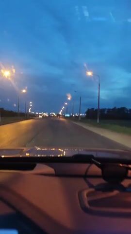 The video shows the view from inside a moving car, looking out over the dashboard and the road ahead. It is early evening, and the sky is a deep blue with wispy clouds. The road stretches out straight, lined with evenly spaced streetlights that cast a yellow glow on the asphalt. There are few other vehicles visible on the road. The hood of the car is visible at the bottom of the frame, reflecting the streetlights.