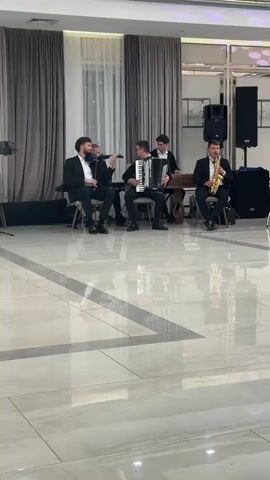 The video shows a band playing music in a well-lit indoor venue. There are five men on stage, dressed in dark suits. The man on the far left is playing the violin. The man next to him is playing an accordion. The man behind the accordion player is playing a guitar or similar string instrument. The man on the far right is playing a saxophone. The stage is set against a backdrop of draped curtains in shades of beige and brown, with a white sheer curtain in the center. The floor is polished and reflective, showing the lights from the ceiling. There are speakers and other audio equipment visible on the stage and to the right. The overall atmosphere appears to be a performance or event.
