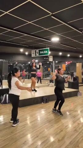 In a brightly lit gym with reflective walls, three women participate in a dance class. The woman in the center is on a raised platform and wears a black crop top and black shorts. The woman on the left wears a white t-shirt and black leggings. The woman on the right wears a black t-shirt and black leggings, with an elaborate design on the back. They all face the reflective walls and perform the same dance moves, including arm raises and torso twists. A green "SALIDA" sign hangs from the ceiling, indicating the exit. Personal items like bags and water bottles are on the floor. Other people can be seen in the background.