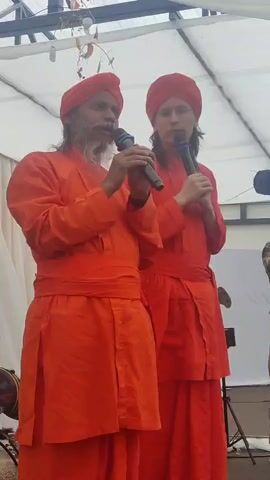 Two individuals are standing next to each other. The person on the left has a long grey beard and is wearing an orange robe and a red turban. They are holding a black microphone. The person on the right is wearing an orange robe and a red turban and is also holding a black microphone. They are both looking towards the left. The background appears to be an outdoor event with a white tent or canopy overhead.