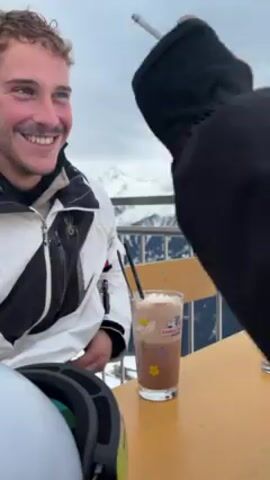 The video shows two people sitting at a table outside a building, likely a ski resort. The person on the left is a young man wearing a white and black ski jacket with curly brown hair and a mustache. He is smiling and holding a cigarette. The other person is mostly out of frame, but you can see they are wearing black ski gloves and have a white ski helmet on the table. On the table there is also a glass of hot chocolate with whipped cream and two black straws, and an empty glass. In the background are snow-covered mountains and a cloudy sky. The video ends with the view tilting down to the person wearing the ski gloves.
