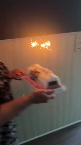 A woman with blonde hair is holding a rectangular cake with lit sparklers on it. The cake is decorated with white frosting, pink frosting on the sides, and sprinkles. The words "Happy Birthday Keith" are written on the cake in dark lettering. The woman is wearing a black and white patterned off-the-shoulder top. She is walking towards a sliding glass door that leads to an outdoor patio. A dog can be seen in the background on the patio. The scene appears to be a birthday celebration.
