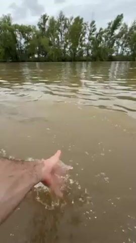 A man's arm and hand are immersed in a body of muddy water, repeatedly scooping and splashing the water. The water is slightly choppy. In the background, a line of trees borders the waterway under a cloudy sky.