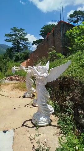 The video opens with a close-up of two white angel statues, one holding a trumpet. The camera then pans up to reveal a bright blue sky with fluffy white clouds. Beyond the statues, a stone wall runs along a hillside, with lush green vegetation and trees. A man in an orange shirt is visible working on top of the wall. Further in the distance, mountains are visible. The camera then moves to the left, revealing a building with a red tiled roof and brown wooden walls, featuring a large star-shaped emblem on the front. Evergreen trees are scattered around the building. The video continues to pan, showing more of the building and the surrounding greenery against the backdrop of the mountains and the blue sky. The sun is shining brightly throughout the video.