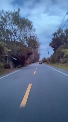 The video shows a view from a moving vehicle on a paved road with yellow lane markings. Trees and vegetation line both sides of the road. The sky is cloudy and appears to be dusk or early morning. In the distance, a yellow car is visible. A motorcycle passes the camera on the right side of the road. Later, two motorcycles are seen on the side of the road on the right. The motion blur indicates the camera is in motion.