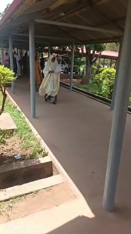 This video shows a group of people walking along an covered walkway. The subjects are primarily women dressed in traditional Islamic attire, with several wearing hijabs and flowing robes in various colors like yellow, white, and brown. One woman in the foreground is wearing a light yellow robe and holding a piece of paper. Another woman in a mustard-yellow robe walks further back. Several individuals in white uniforms, possibly nurses or medical students, are visible in the background.

The setting appears to be an outdoor walkway connecting buildings, likely a hospital or educational institution, given the presence of uniformed individuals. Greenery and trees are visible on the left side of the walkway, suggesting a daytime scene, possibly late morning or early afternoon given the lighting and shadows. The walkway itself is paved with concrete and supported by pillars. The overall atmosphere seems calm and orderly as the group proceeds along the path.

The visible text at the bottom of the screen reads "TECNO CAMON 40 Pro," indicating the device used to record the video. There are no overt signs of distress or urgency, suggesting a routine movement or procession. The environment seems well-maintained. The location context of Tudun Nupawa, Nigeria, combined with the attire of the individuals, suggests a cultural and regional setting.