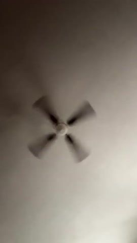 The video shows a person, likely male, filmed from a low angle, looking up at a ceiling fan. The person's face is visible in the foreground, with a surprised or playful expression. They appear to be in an indoor setting, as indicated by the ceiling fan. The lighting is dim, suggesting it could be nighttime or a room with minimal natural light. The person's upper body is partially visible, and they seem to be wearing a light-colored shirt. The ceiling fan is in motion, with its blades blurred due to the rotation. The video is short, with no discernible dialogue or music. The person's pose and the camera angle create a dynamic and somewhat disorienting effect. No text is visible in the video. The location context of Sesto Calende, Italy, does not provide any specific visual cues within the video itself.