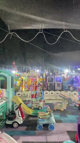The video shows a children's play area at night. It is covered by a dark tarp or canopy, with strings of colorful LED lights running across it. The play area is filled with various toys and equipment, including small ride-on cars, a playhouse with a slide, a swing set, and other colorful play structures. Several arcade games are visible in the background. The floor is covered with interlocking foam mats in shades of purple and gray. The overall impression is of a dimly lit, festive indoor playground.