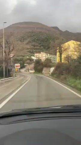 A car drives on a gray road on a cloudy day. In the distance, there is a low, tree-covered mountain. A large billboard advertising burgers sits near a junction. The road has white lane markings, and the windshield of the car is visible.