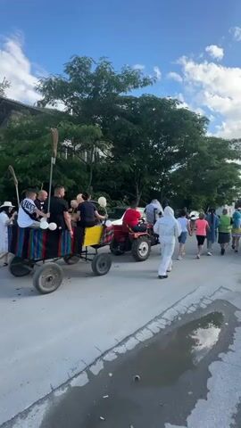 A group of people are walking down a street next to two tractors pulling trailers. One trailer has multiple people sitting on it, with white balloons and brooms visible. The other tractor is red and has a person sitting on it. The people walking are wearing various clothing, with one person in a white full-body suit. The street is paved, and there's a muddy puddle on the right side. Trees and buildings line the street. The sky is blue with some clouds.